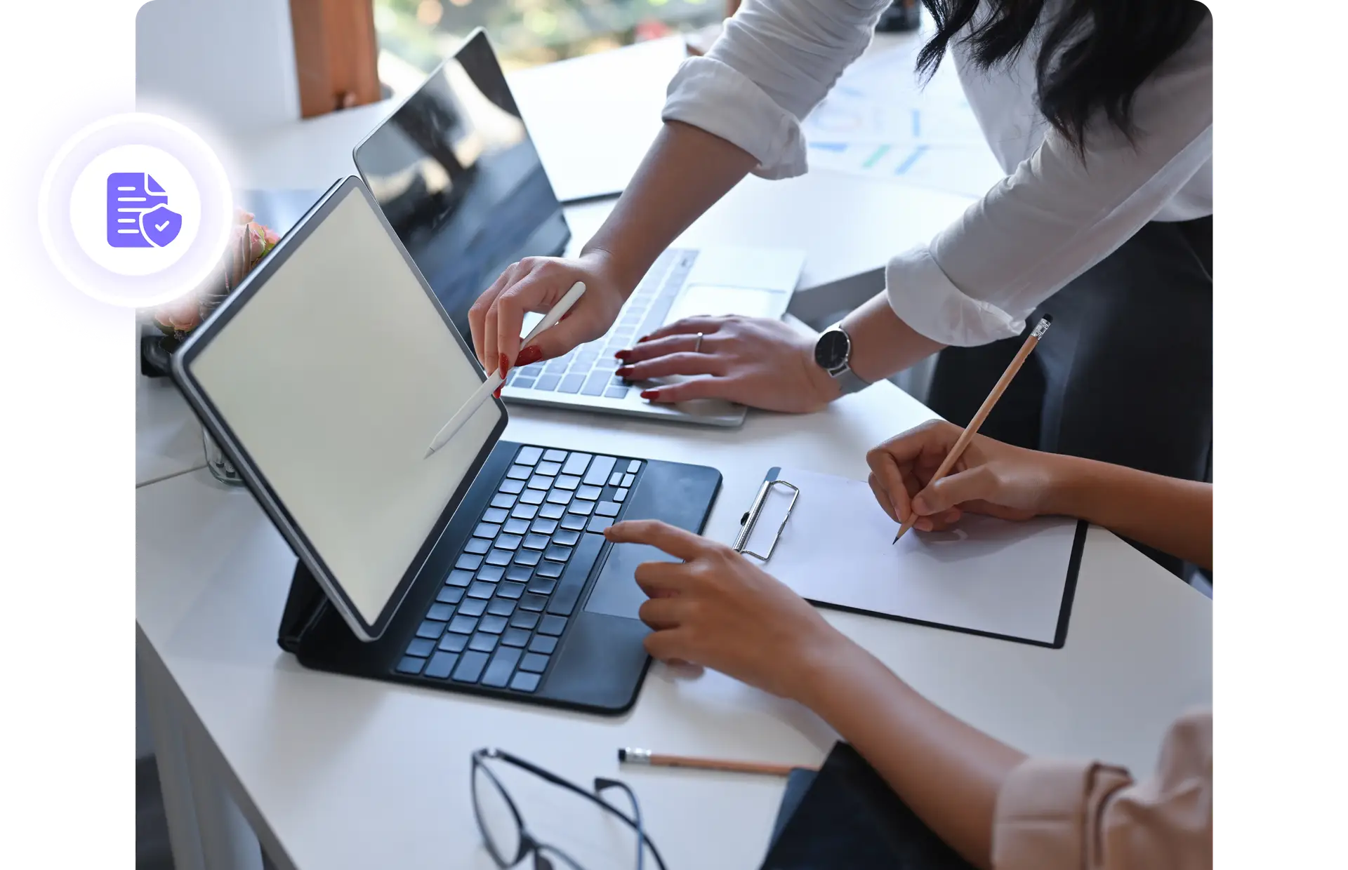 Two persons standing by a desk and pointing with a pen on a laptop screen at the same time as the other take notes on a paper.